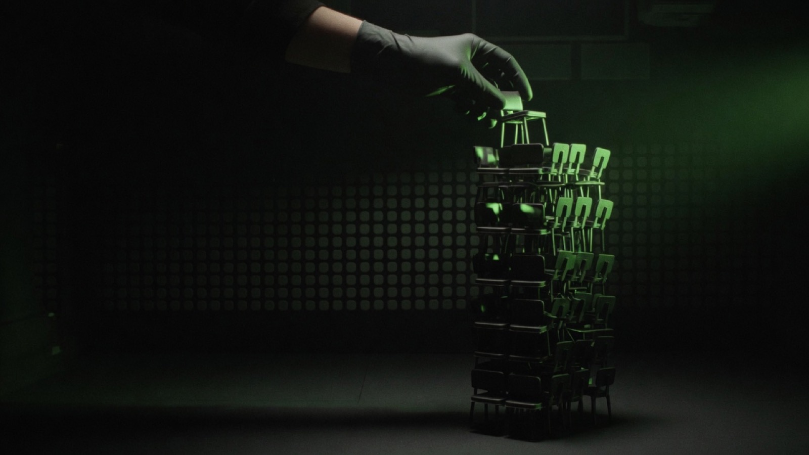 A gloved hand sets a tiny black chair on top of a tall stack of identical chairs in a dark room lit green.