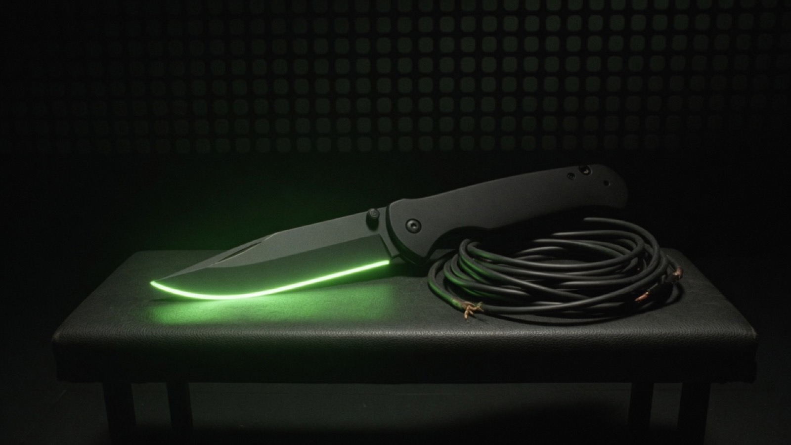 An open matte pocket knife lies on a dark bench, its blade edged in green light, beside a coil of cut cable.