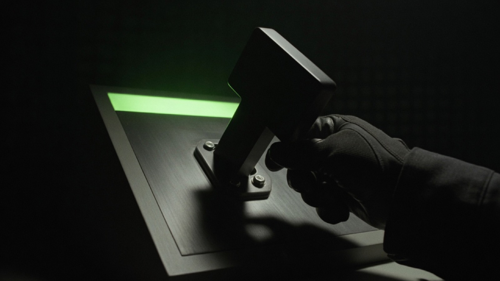 A gloved hand bolts an oversized black key onto the bare face of a dark machine, with a green light in the panel.