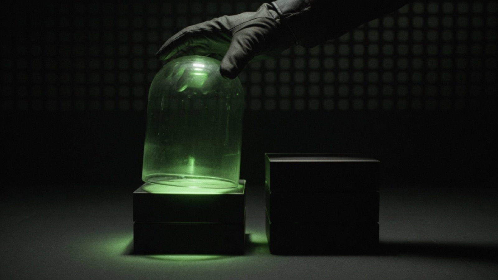 A gloved hand lowers a glass bell jar over a short stack of black boxes beside a taller uncovered stack, the covered stack lit green.