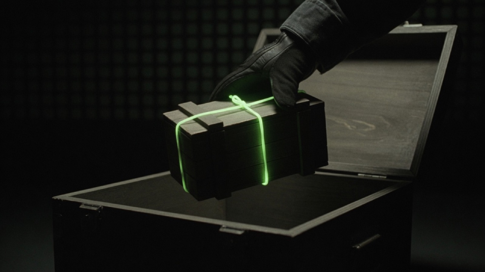 A gloved hand lowers a small closed crate tied with a glowing green cord into a larger open crate.