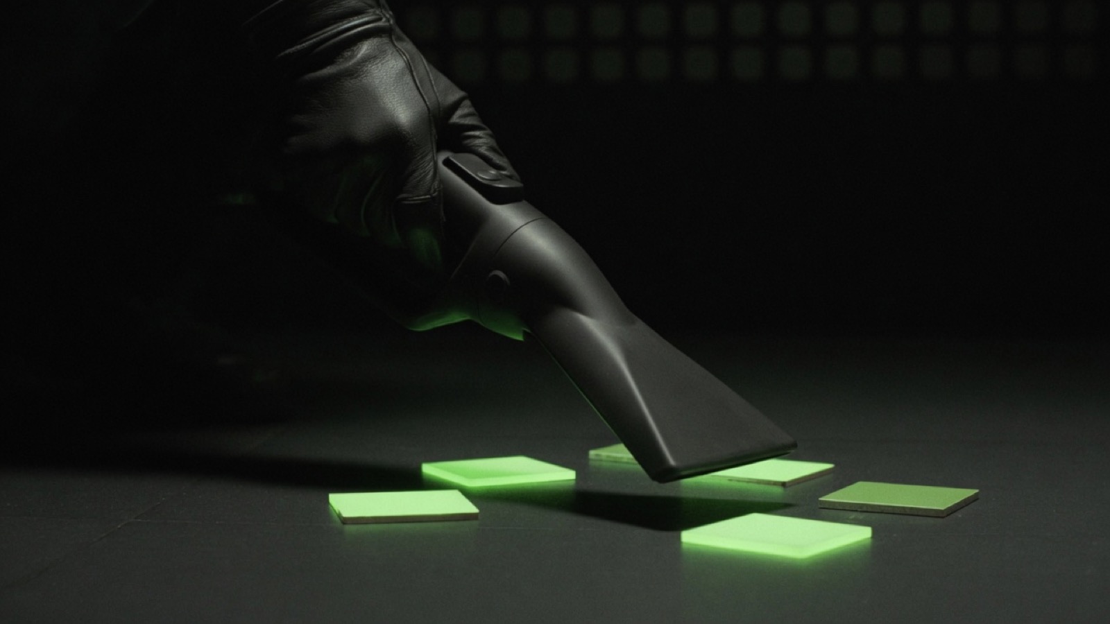 A gloved hand holds a vacuum nozzle over glowing green squares on a dark floor.