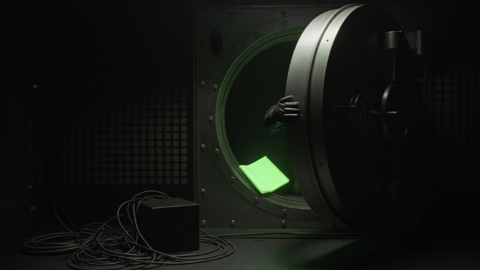 A gloved hand holds a round vault door nearly shut on a glowing notebook, with cables and a small black box left on the floor outside.