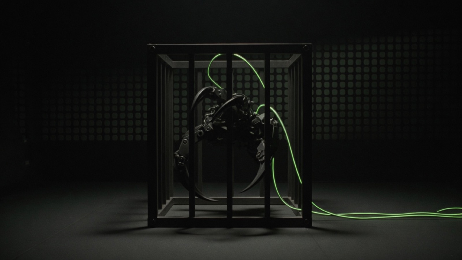 A dark mechanical claw is locked inside a small barred cage, with green cables running out across the floor.
