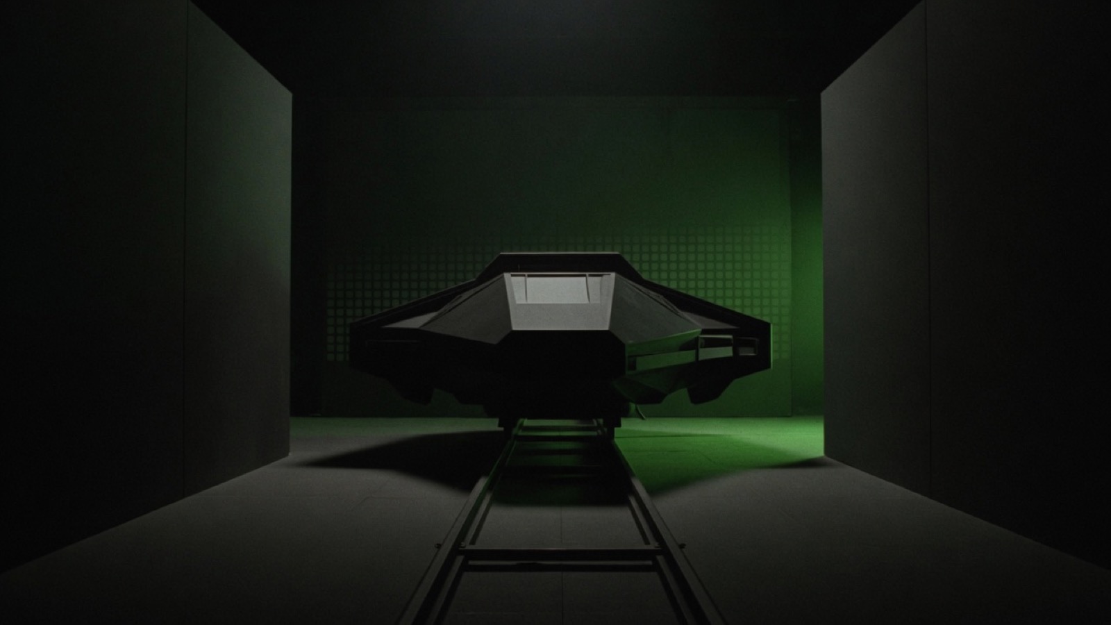 A matte-black spacecraft sits on a straight rail between tall side walls, facing a solid end wall.