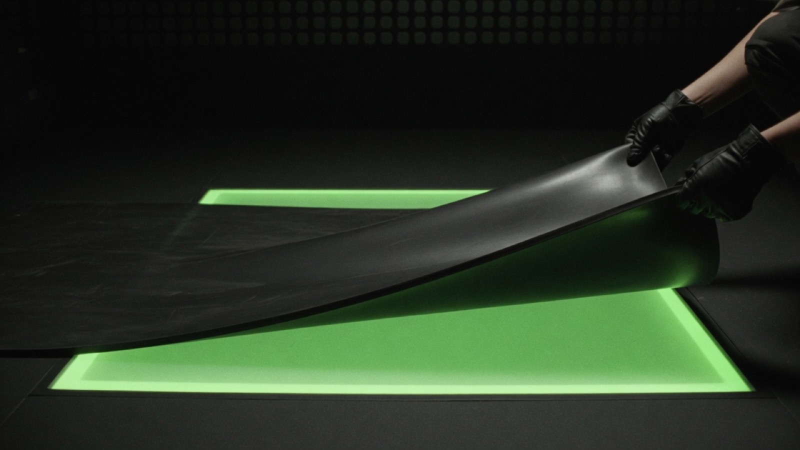Gloved hands pull a black sheet across a glowing green rectangle set into a dark floor.