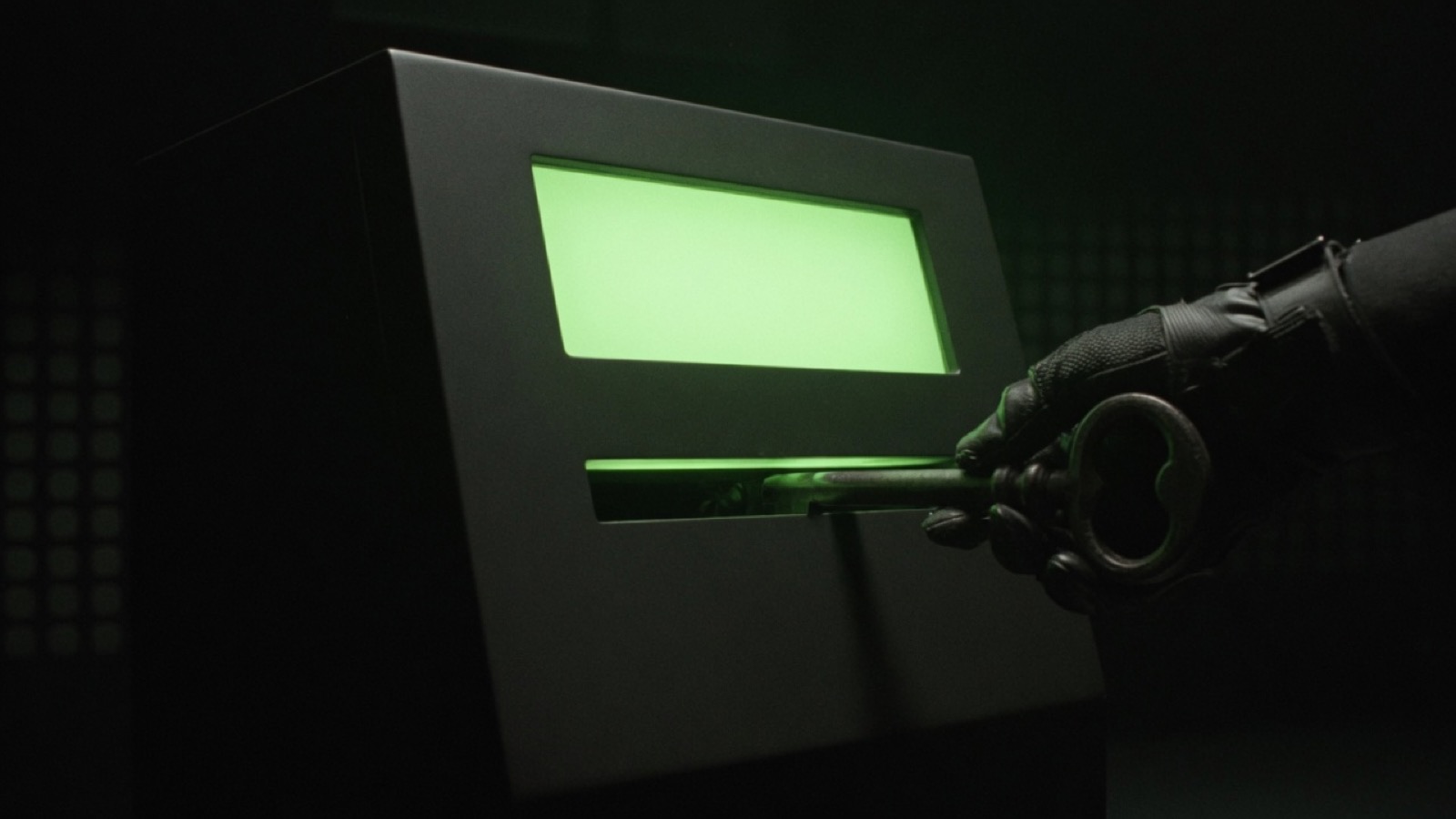 A gloved hand slides an oversized iron key into the slot of a matte black machine whose face is a blank green rectangle.